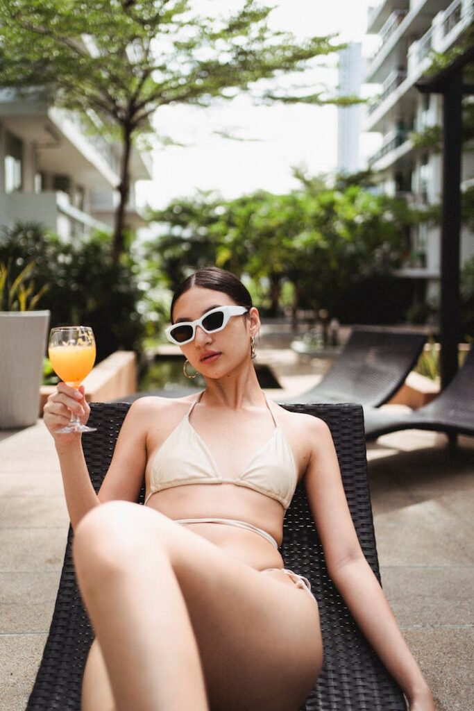 Alluring young slim ethnic woman in bikini and sunglasses chilling on sunbed at poolside of modern hotel and enjoying refreshing juice on holiday